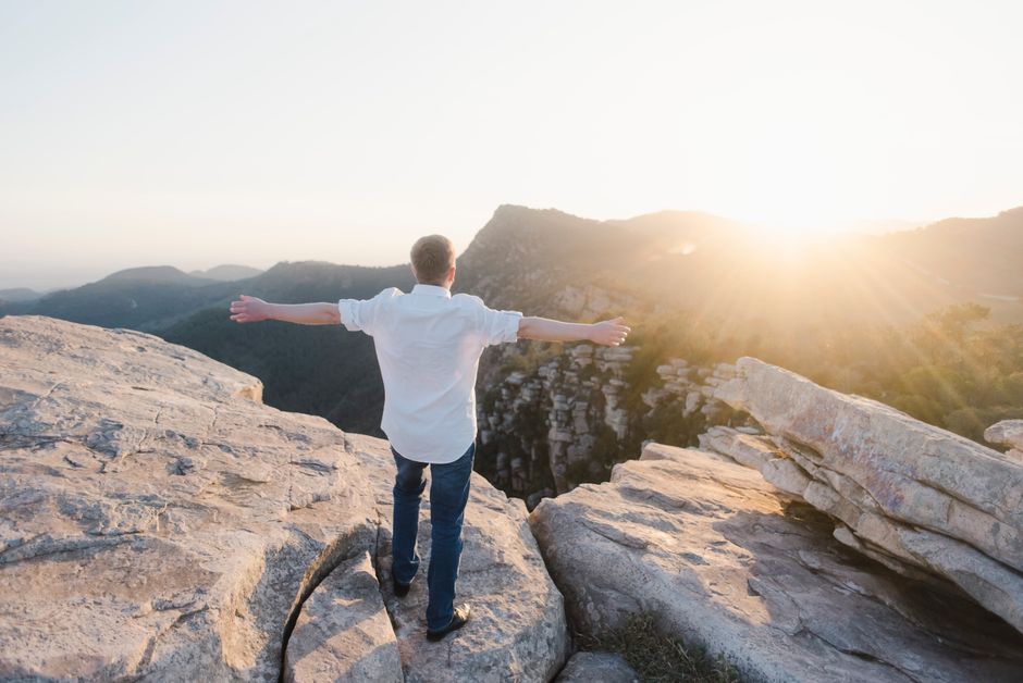 A man stands on a rocky cliff with arms outstretched, embracing the sunset in the mountains.