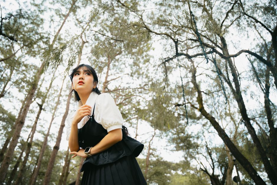 A young woman in a forest, gazing upwards, wearing a black dress and white shirt.