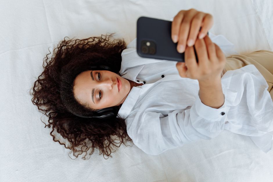 Young woman lying on bed with headphones and smartphone, enjoying music.