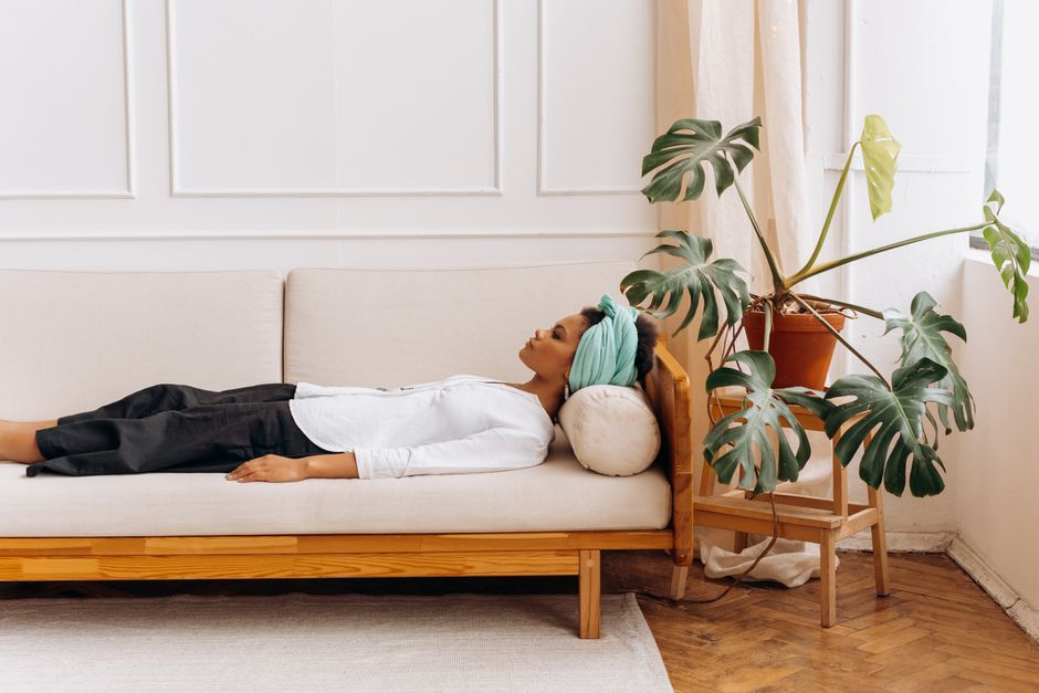 A woman relaxes on a sofa, wearing a headscarf, indoors with a large plant nearby.