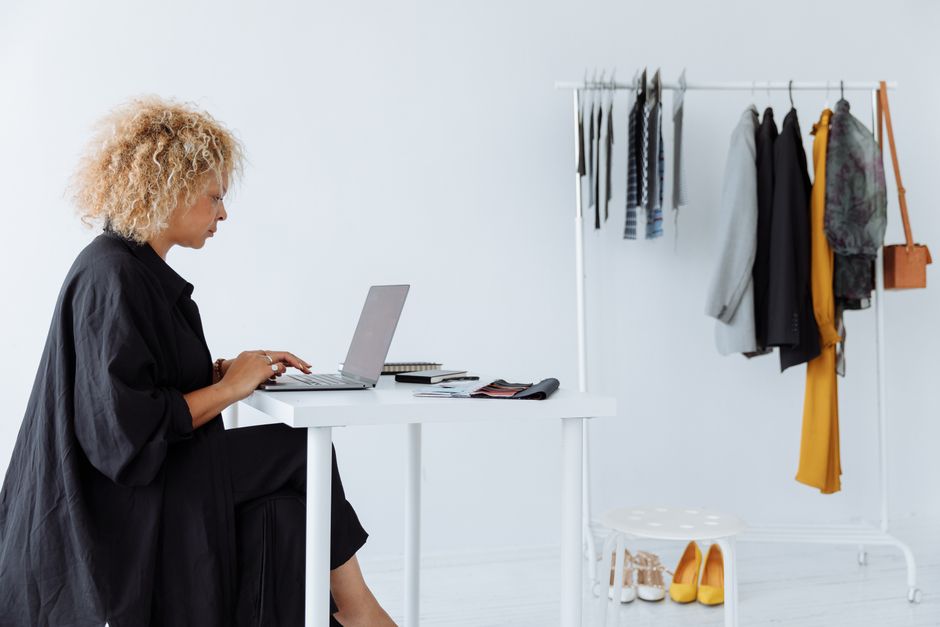 Businesswoman working on a laptop at a white desk with a clothing rack in a bright, minimalist office.