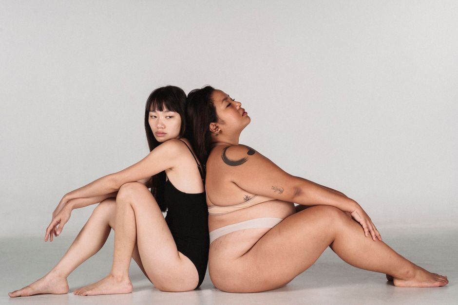 Side view of Asian women in underwear sitting back to back in studio against grey background
