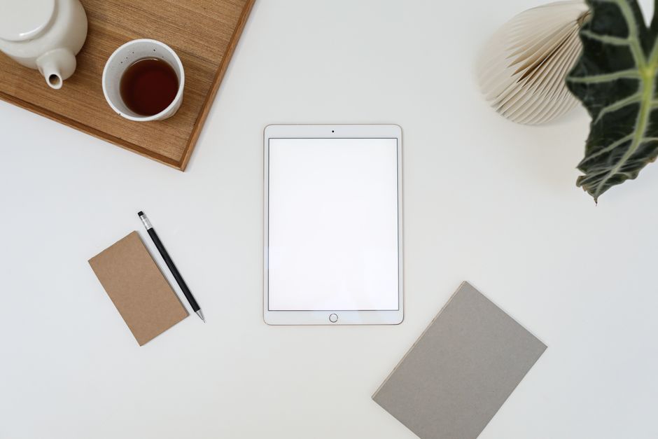 Top view of tablet with empty screen placed near cup of tea and notebook on white table