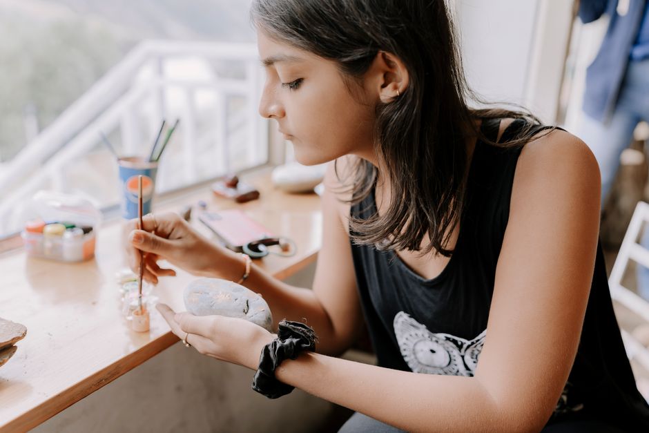 A young woman focused on painting stones by a sun-drenched window in an art studio.