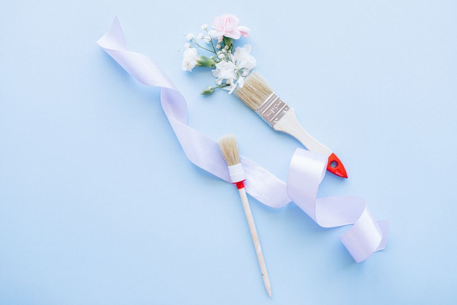 Two paint brushes with ribbon and flowers on a pastel blue background, ideal for DIY and decor themes.