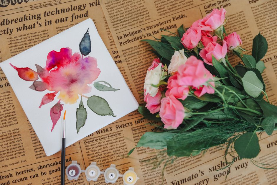 Vibrant watercolor painting of a flower alongside pink roses on newspaper.
