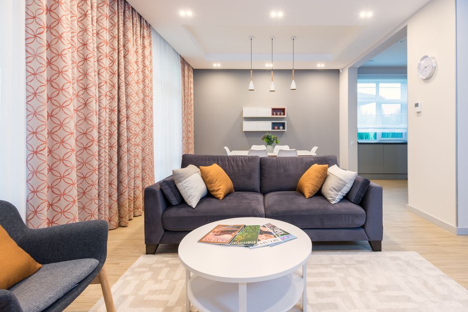 Comfortable sofa and armchair placed on carpet near side table with magazines and kitchen table in stylish spacious apartment with window