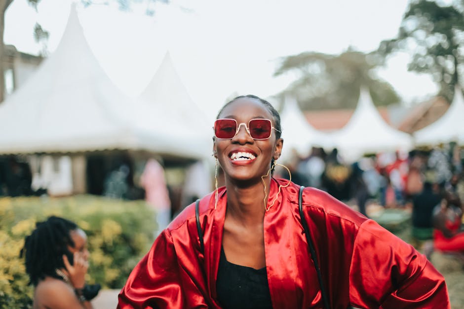 A woman in sunglasses and red attire smiling at an outdoor festival with tents.