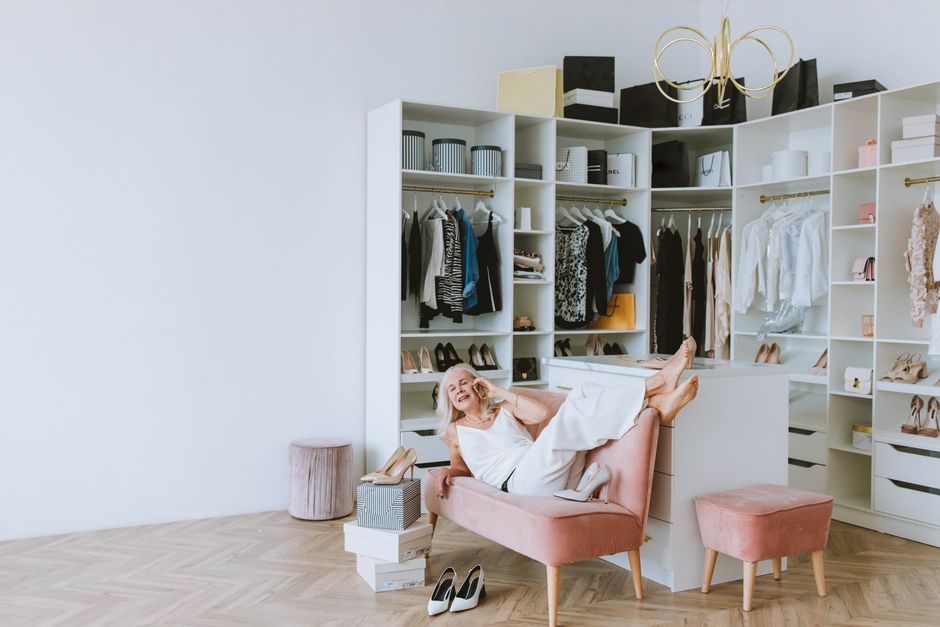 Senior woman lounging in a stylish dressing room with clothes, shoes, and shopping bags.