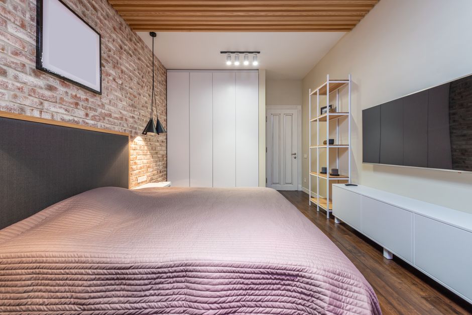Bed with cover under empty panel on brick wall against television with black screen above cabinet at home