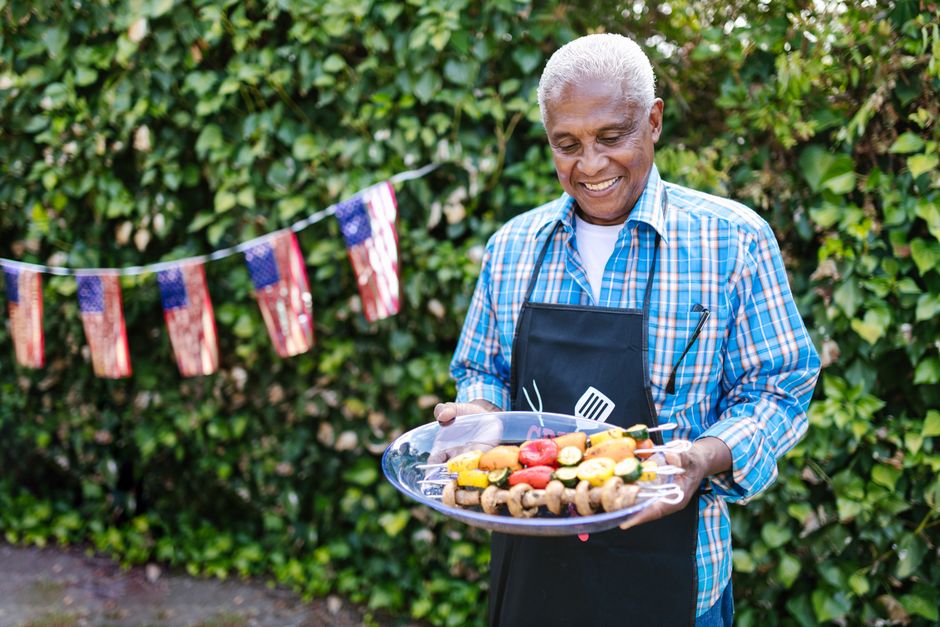 Senior man holding skewers at 4th of July barbecue with USA flags in garden.