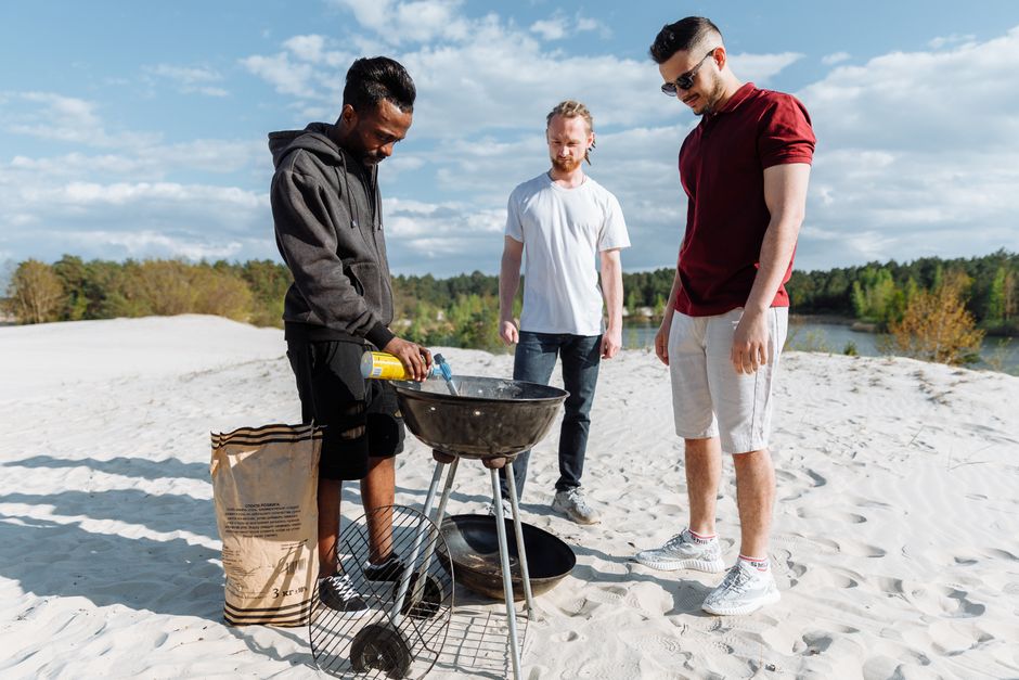 Three men preparing a grill on a sandy beach with clear sky, enjoying outdoor BBQ.